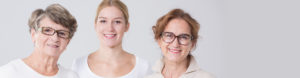 Advanced Skin + Body Aesthetics Med spa, Botox, Facials and more! Three women of different ages, smiling at the camera, standing side by side against a light gray background at Med Spa Lincoln NE. They wear casual white tops and glasses.