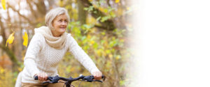 Senior woman in a white sweater rides a bicycle in a sunny autumn park, surrounded by fall foliage near Med Spa Lincoln NE.