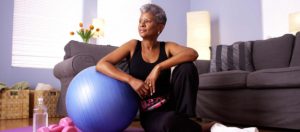 A senior woman with short gray hair sitting on a large blue exercise ball in a Med Spa Lincoln NE, dressed in workout attire, resting her arm and looking thoughtfully to the side.