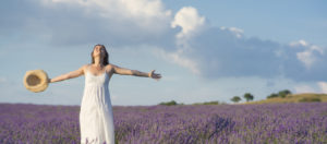 A woman in a white dress and holding a hat, arms outstretched, joyfully stands in a blooming lavender field under a blue sky with clouds, reminiscent of the serene ambiance at Med Spa
