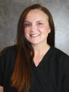 A professional portrait of a smiling woman with long brown hair, wearing a black medical scrub, standing against a mottled gray background.
