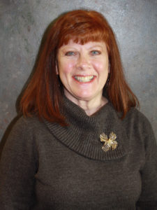 A woman with shoulder-length red hair and a broad smile, wearing a gray turtleneck and a gold brooch, posed against a mottled gray backdrop.