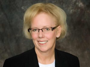 A professional portrait of a smiling middle-aged woman with short blonde hair, wearing glasses, pearl earrings, and a black blazer against a mottled gray background.