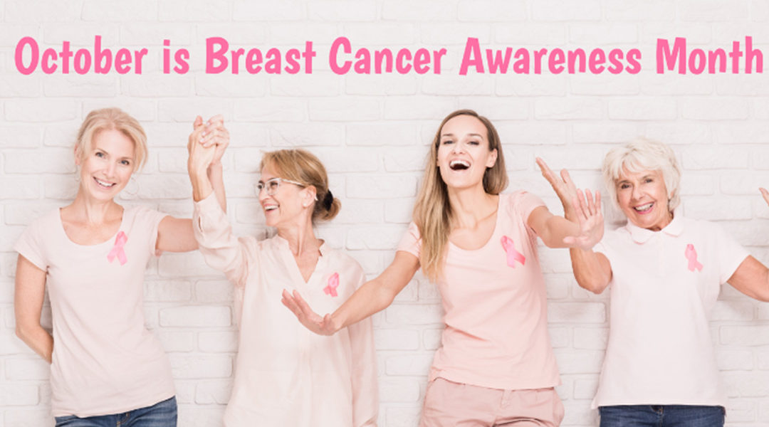 Four women smiling and raising their hands joyfully, wearing pink t-shirts with breast cancer awareness ribbons, standing against a white brick wall with text "october is breast cancer awareness month.