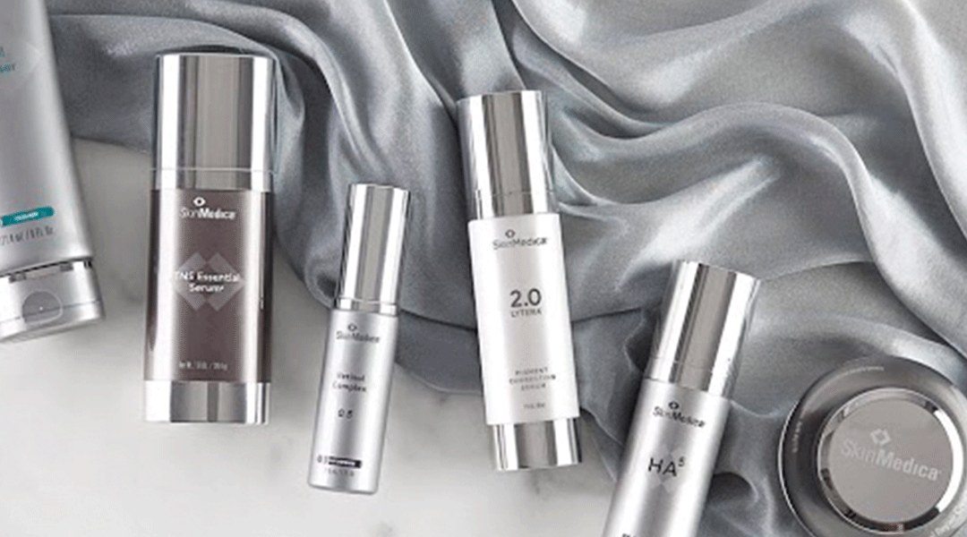 A collection of six skinmedica skincare products arranged on a gray silky fabric. the items include a facial cleanser, serums, and creams in sleek metallic and white packaging.