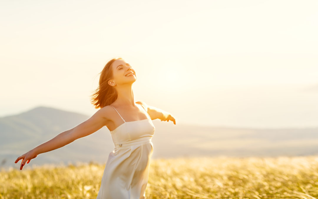Advanced Skin + Body Aesthetics Med spa, Botox, Facials and more! A joyful woman in a white dress with her arms outstretched, standing in a sunlit field against a soft mountain backdrop, embracing the warmth and light of the summer sun.