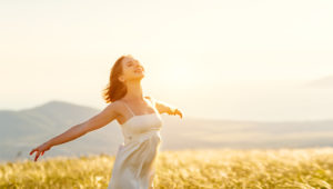 A joyful woman in a white dress with outstretched arms, embracing the sunlight in a golden field with distant mountains in the background.