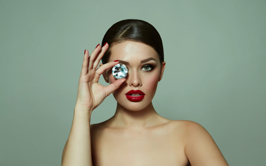 Advanced Skin + Body Aesthetics Med spa, Botox, Facials and more! A woman with elegant makeup, possibly following one of her facials in Lincoln, NE, holds a large diamond in front of her eye, set against a neutral background. Her red lipstick contrasts with her
