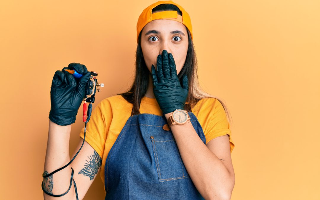 A surprised female tattoo artist with a yellow cap and blue apron holds a tattoo machine, covering her mouth with her gloved hand against a yellow background.