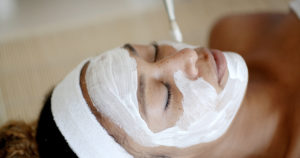 A woman receiving a facial treatment with a white mask applied to her face, lying with her eyes closed and a serene expression.