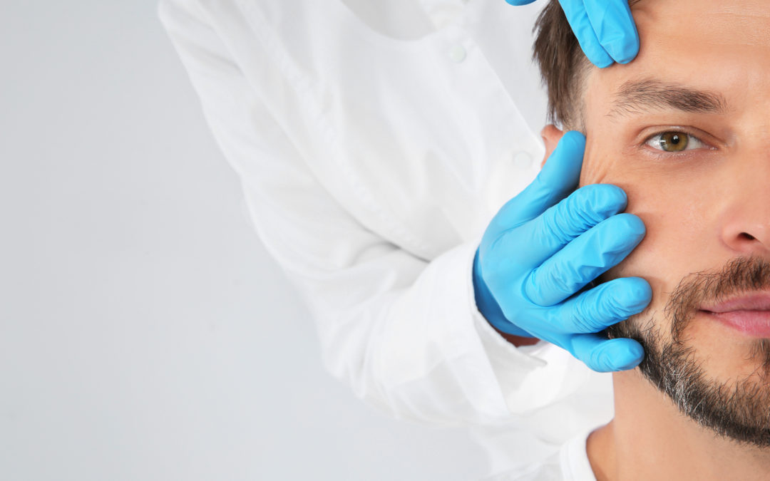 A close-up image of a man's face being examined by a dermatologist wearing blue gloves. only the hands of the doctor are visible. the background is a neutral light gray.