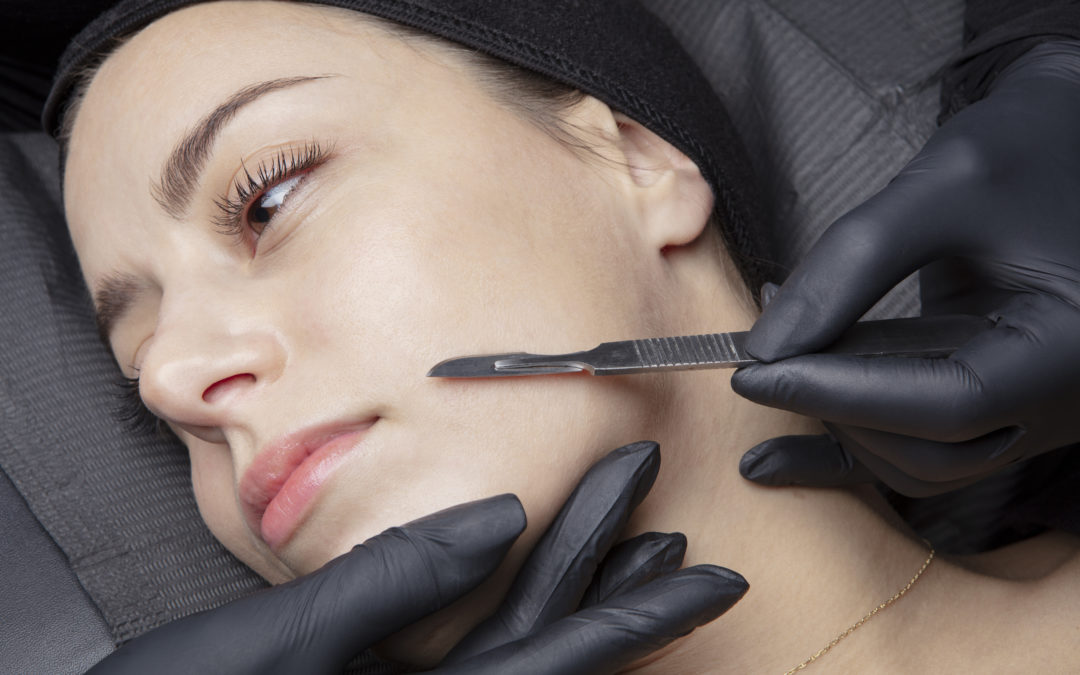 Advanced Skin + Body Aesthetics Med spa, Botox, Facials and more! A woman lying down with her eyes closed, having a dermaplaning procedure done on her face using a scalpel by a professional wearing black gloves.