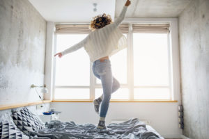 A joyful woman with curly hair jumping on a bed in a sunlit room with Lincoln aesthetics, gesturing triumphantly with her arms raised.