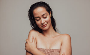 A young woman with wet hair and freckles softly embracing her own shoulder, eyes closed, against a neutral background. she exudes a peaceful and content expression.